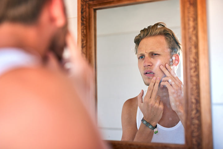 Handsome young man examining his skin in the mirror, looking for blackheads and spots