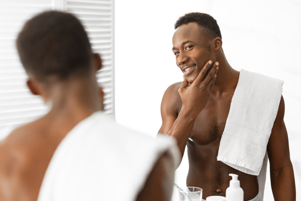 Shaving Black Skin: How to Get a Clean, Smooth Shave – No Razor Bumps, No Dark Spots. Handsome black man looking in the mirror feeling his smooth chin